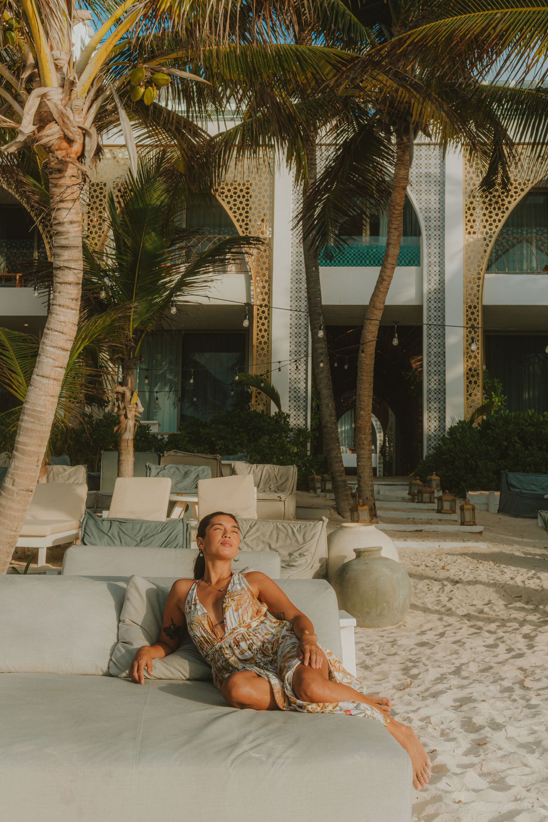 Woman relaxing on a cushioned daybed, beach setting, palm trees, resort building with arches.