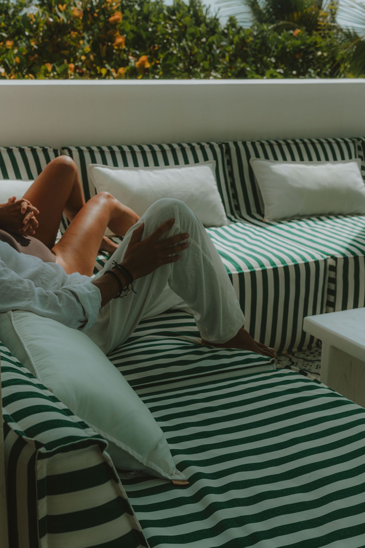 Person reclining on striped green and white outdoor sofa with white pillows; ocean view.