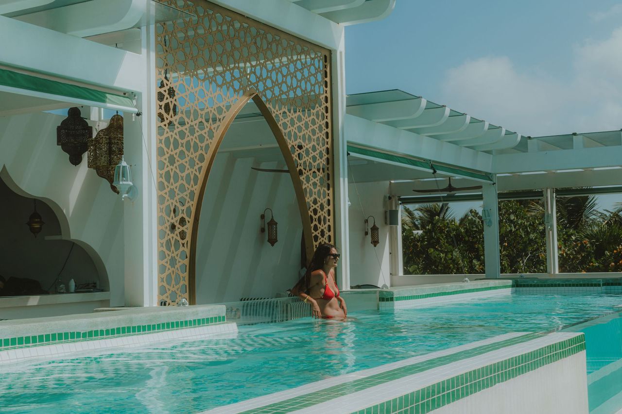 Woman in a red swimsuit relaxes in a turquoise pool under a white and gold archway, bright sunlight.
