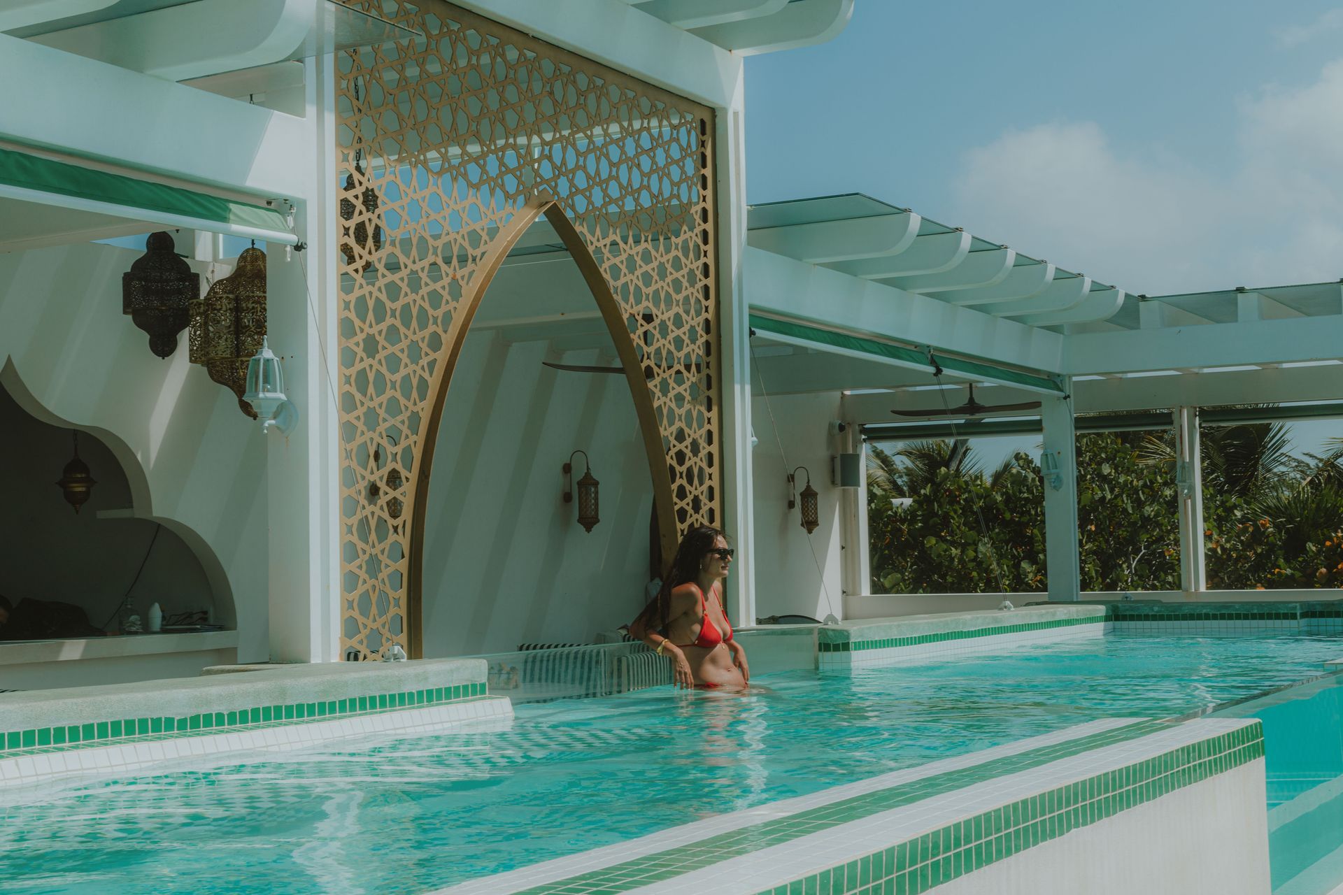 Woman in a red swimsuit relaxes in a turquoise pool under a white and gold archway, bright sunlight.