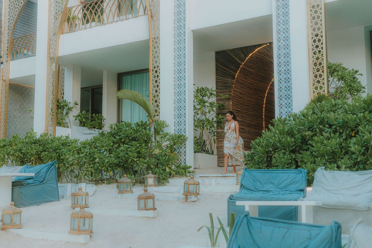 Woman in a dress exits a white building with teal accents and decorative cutouts. Lush greenery surrounds the entrance.