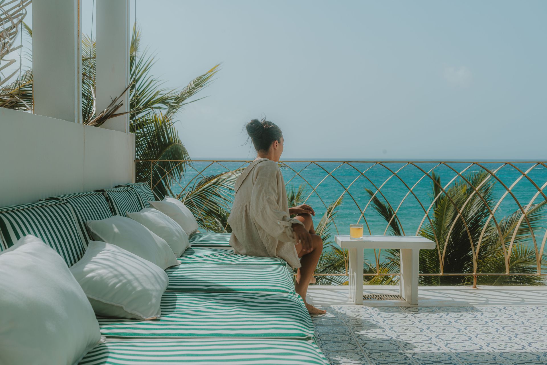 Woman sitting on striped sofa, overlooking the ocean. She holds a drink, the scene is serene and sunny.