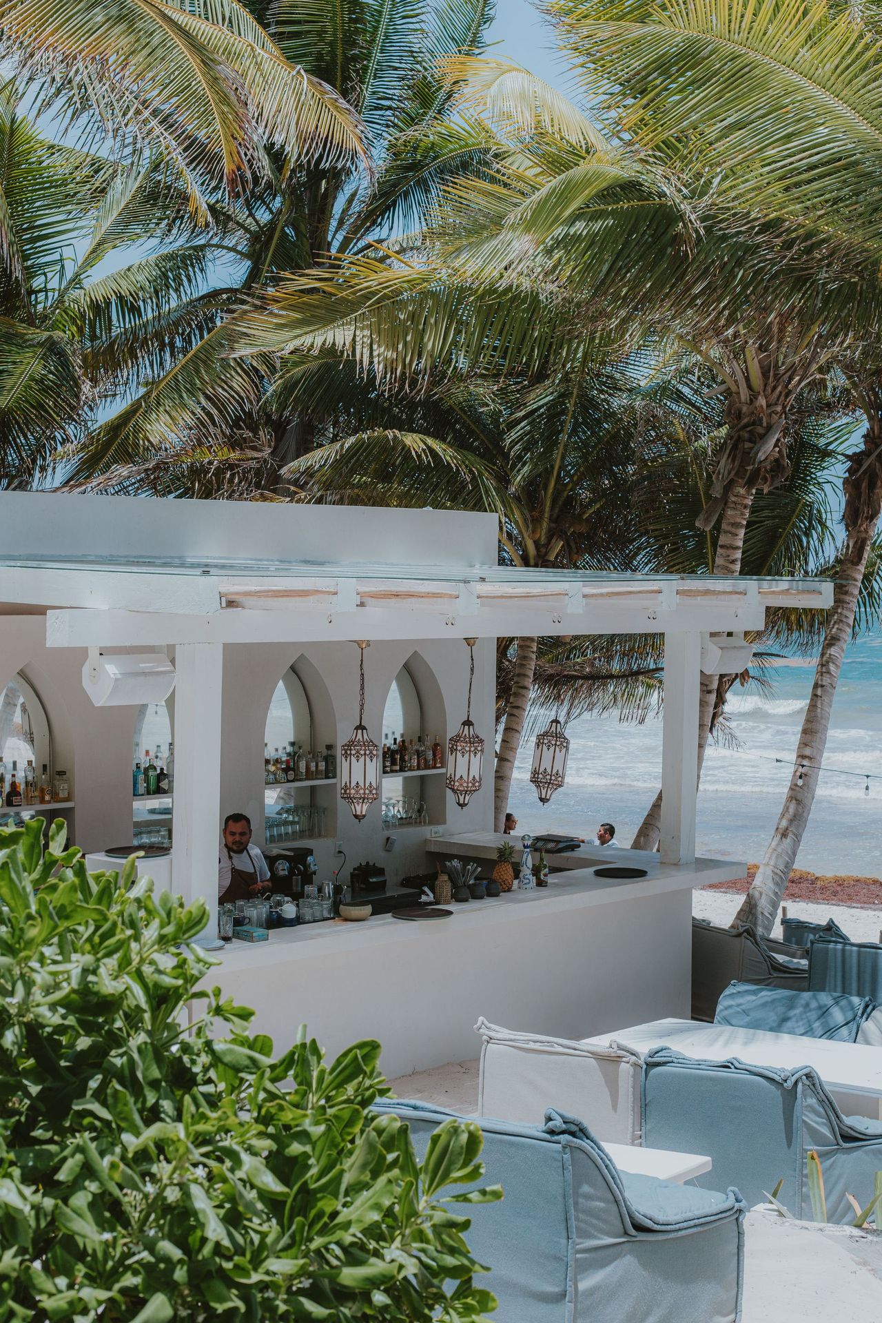 White beachside bar with palm trees, ocean view, and server behind the counter.