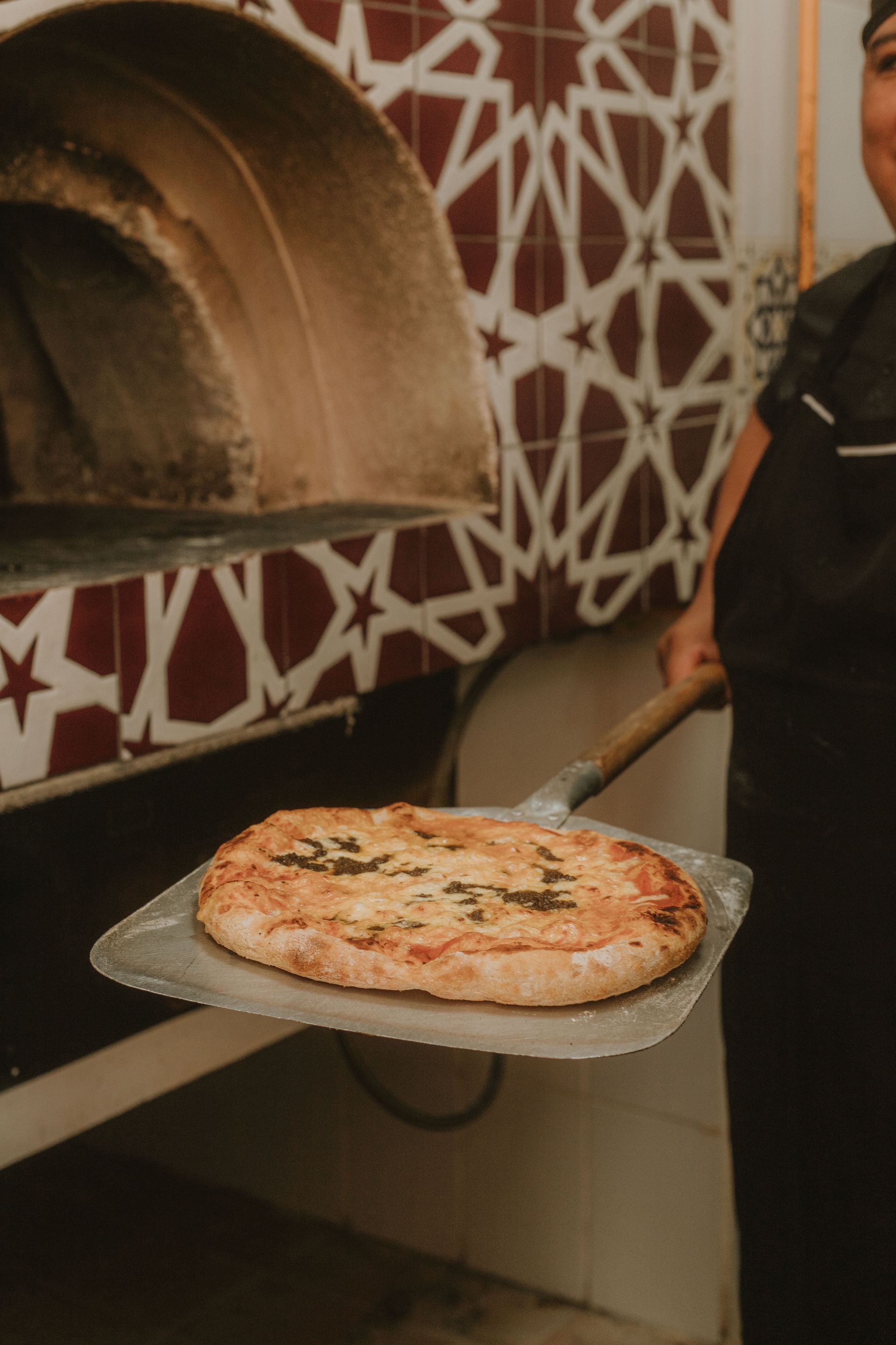 Pizzero sosteniendo una pizza recién horneada en una pala frente a un horno de ladrillos.
