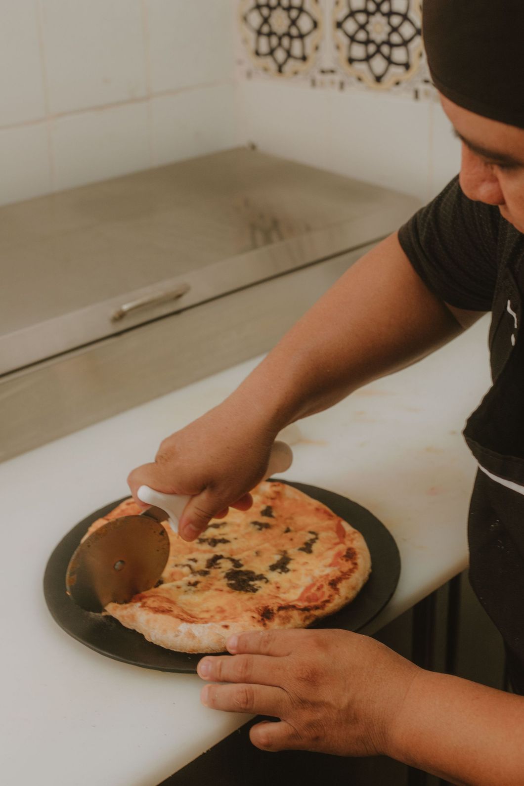 Person cutting a pizza with a wheel cutter on a dark plate, next to a stainless steel appliance.