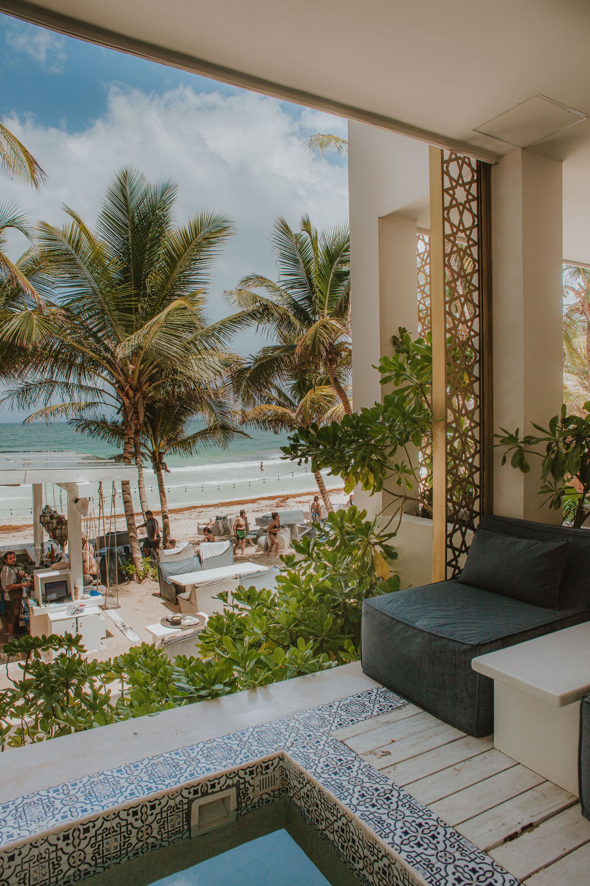 Balcony with beach view, blue mosaic jacuzzi, gray seating, and palm trees.