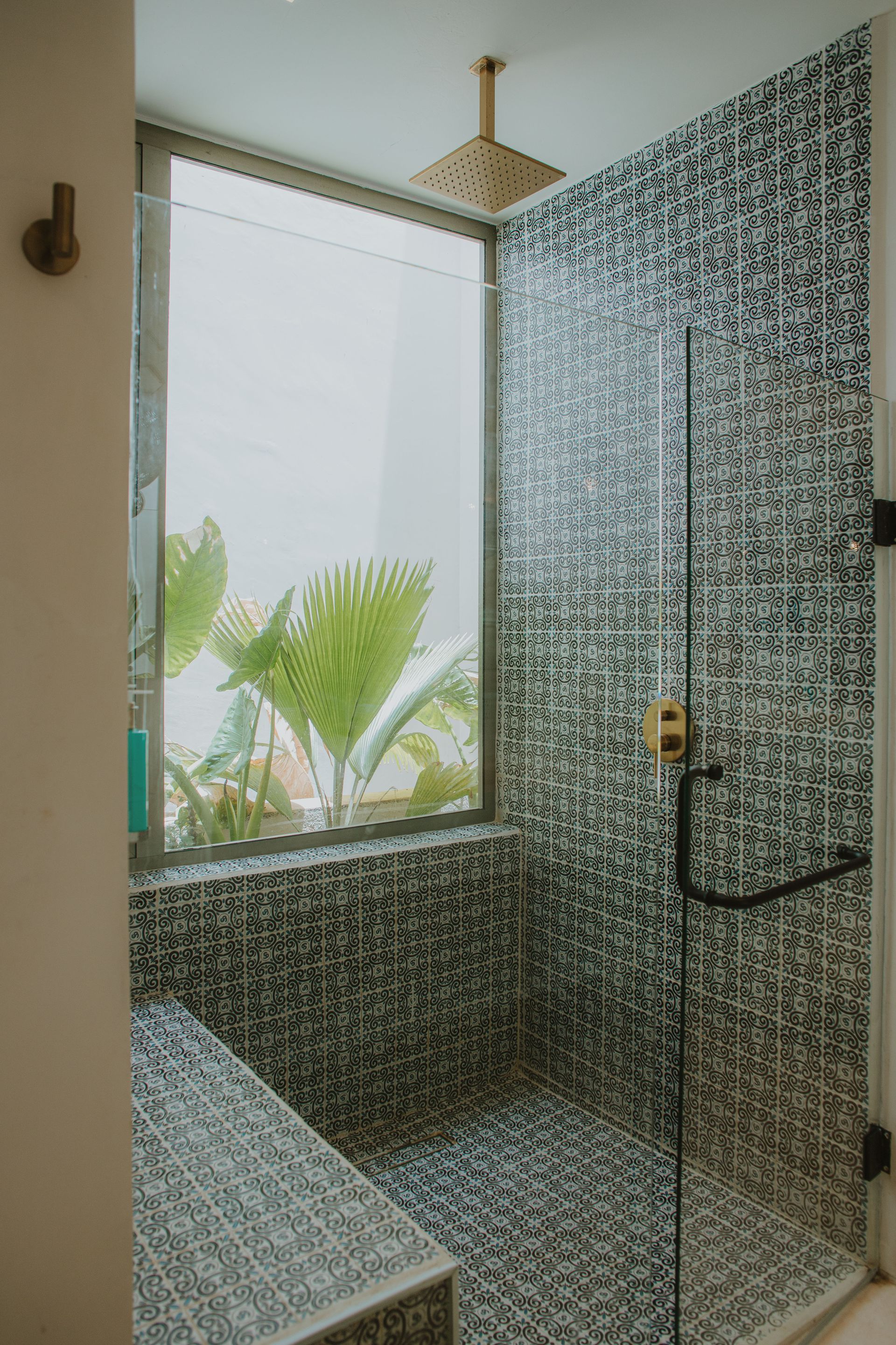 Shower with mosaic tile, glass door, and window overlooking greenery. Gold showerhead, black handle.
