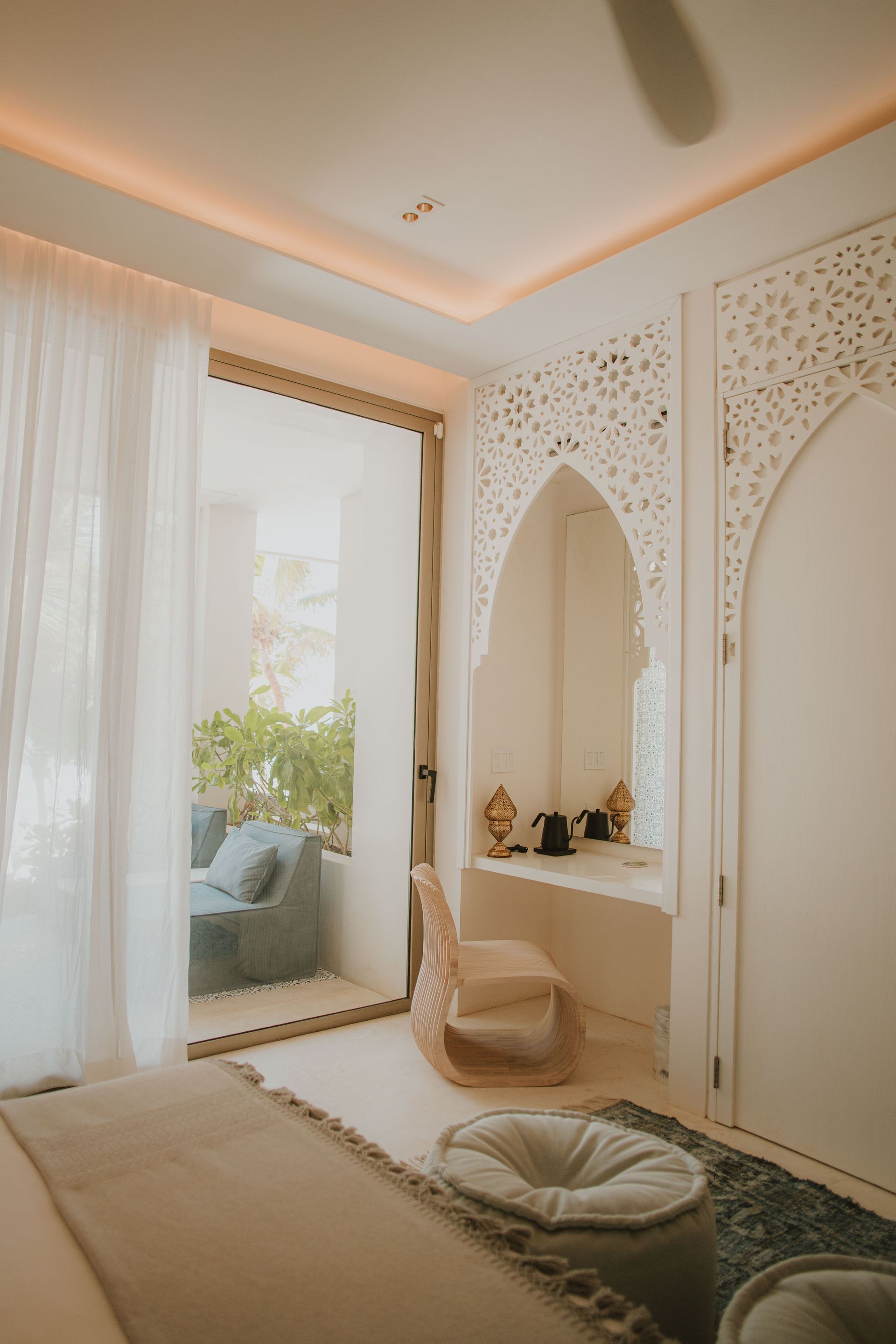 Bedroom with white carved wall, open to a balcony with blue seating. Sunlight streams in.