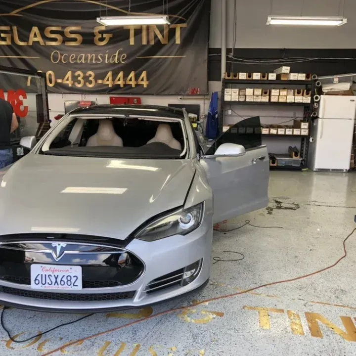 Silver Tesla being worked on at a glass & tint shop; open doors, clear window, Oceanside signage in background.