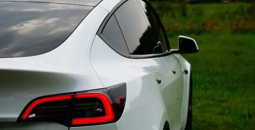 White Tesla car's rear quarter panel and taillight. Dark tinted windows, green grass in the background.