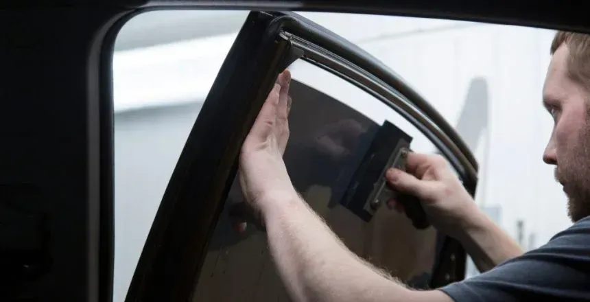 Man applying window tint to a car window with a squeegee inside a vehicle, likely a garage.