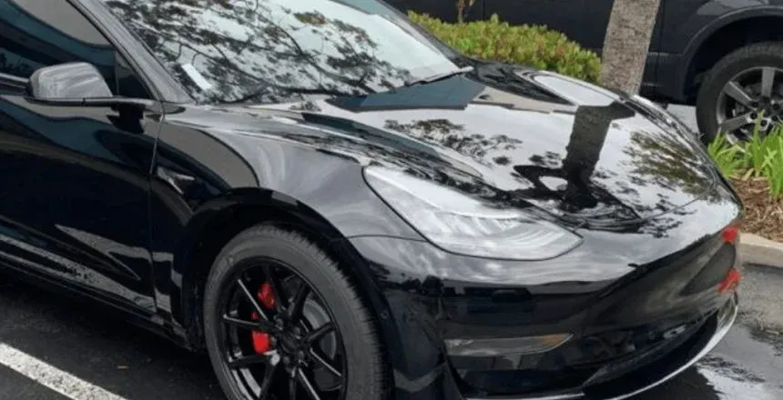 Black Tesla Model 3 parked, shiny, with red brake calipers. Sunlight reflects on the hood.