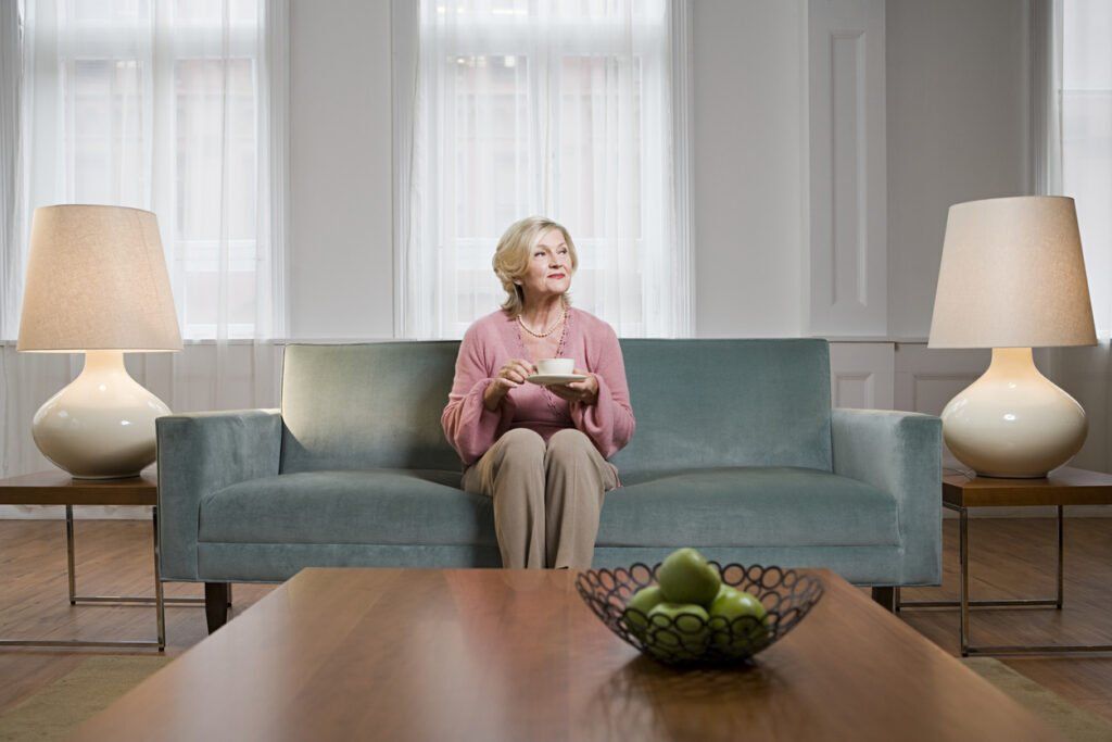 Elderly woman sits on gray couch with a cup of tea