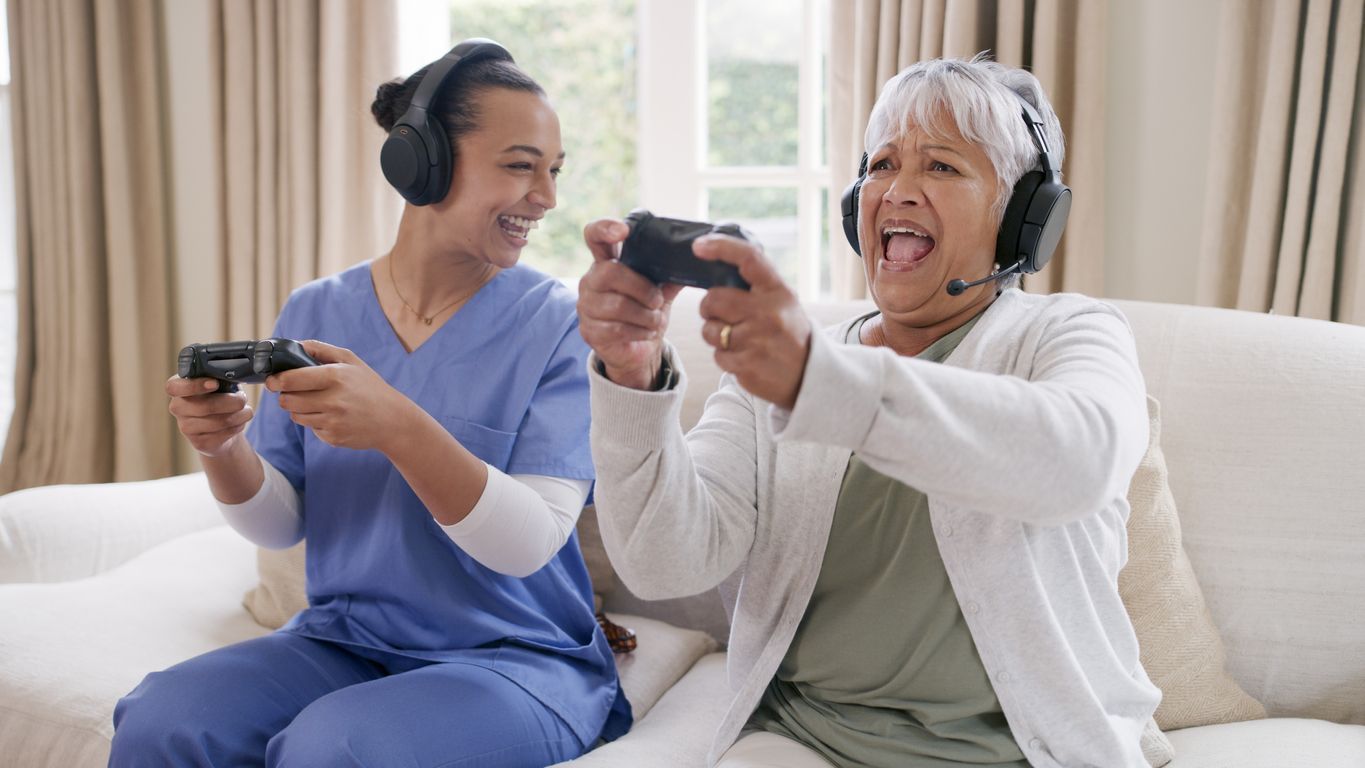 senior woman playing video games with female caretaker on couch. Headsets and controllers.