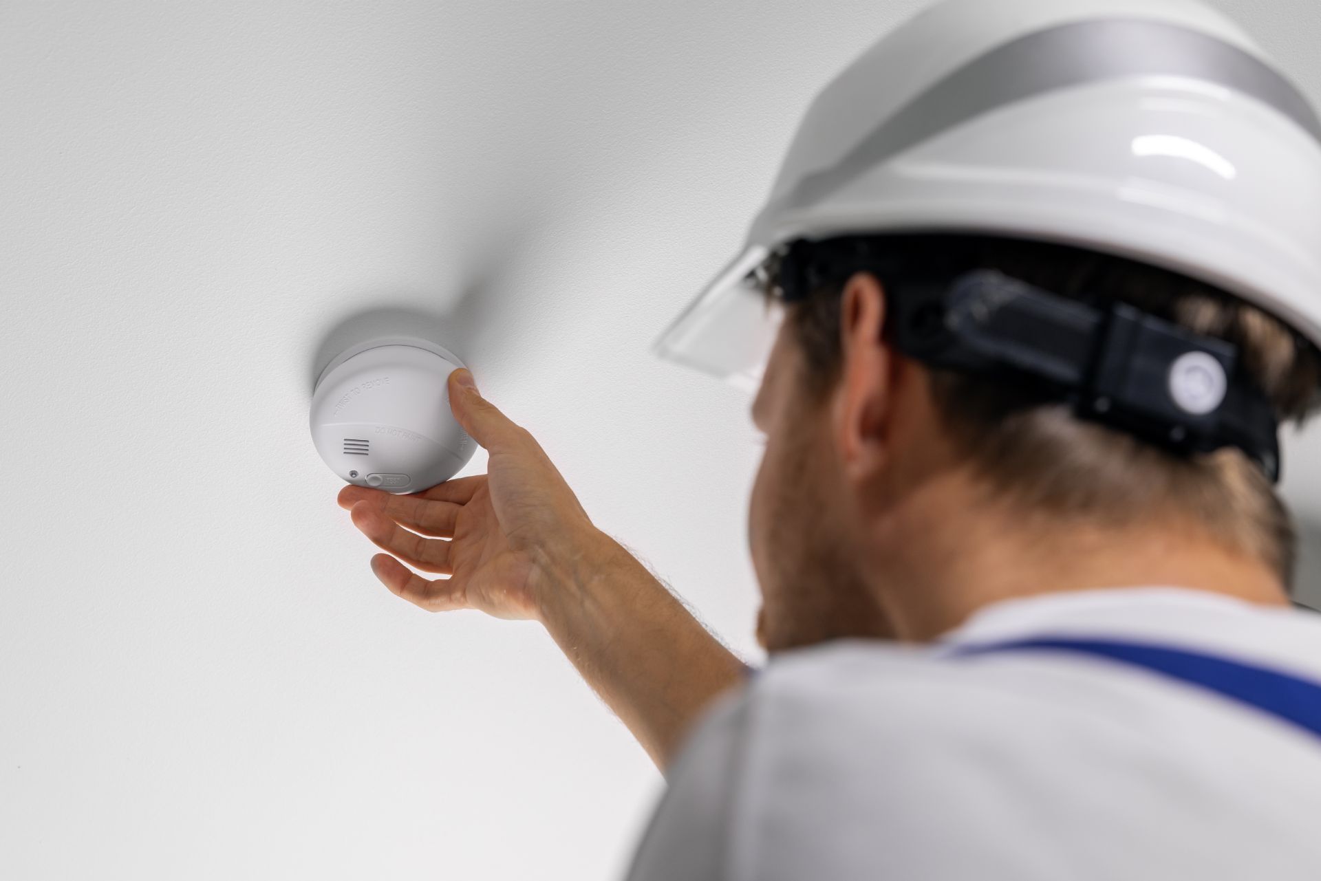 Man in hard hat installs a smoke detector on a white ceiling.