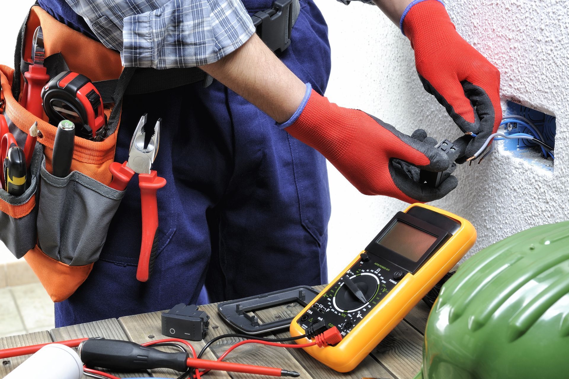 Electrician wearing red gloves working on an outlet with tools and a multimeter on a wooden surface.