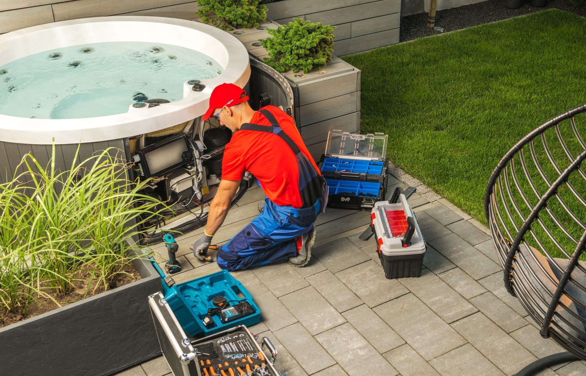 Man in red shirt and blue overalls repairs hot tub outdoors, surrounded by tools.