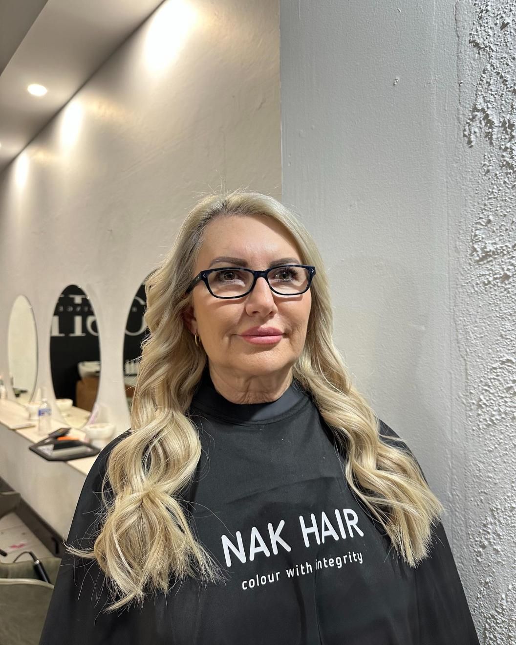 A Woman Wearing Glasses and A Black Apron Is Standing in A Salon — Losh Hair Lounge In Toowoomba City, QLD
