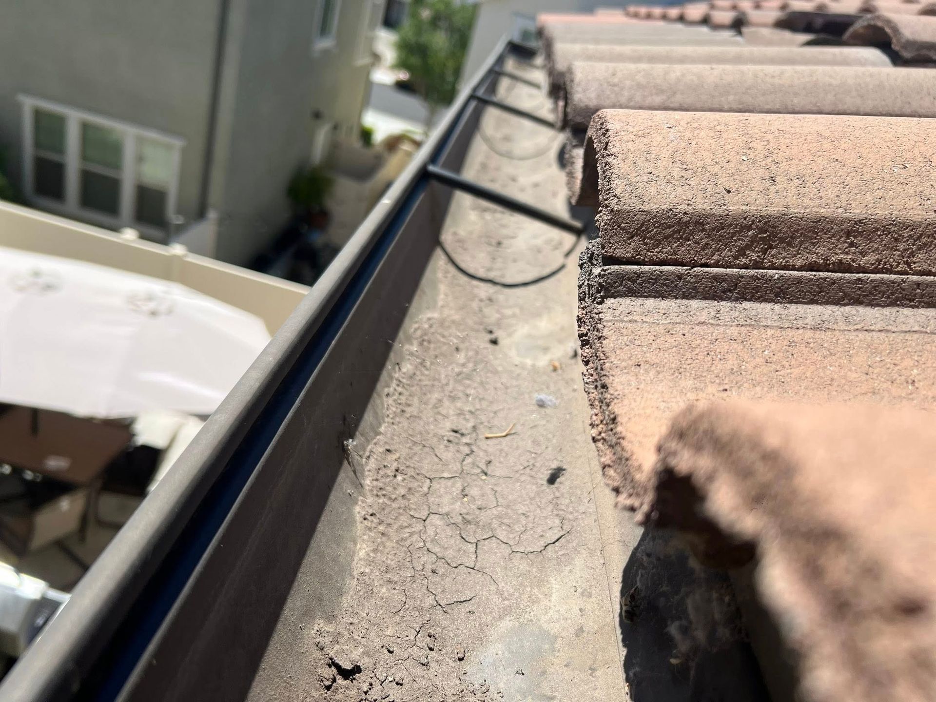 A close-up view of a roof gutter and adjacent clay roof tiles on a sunny day, showing debris and metal support clips.
