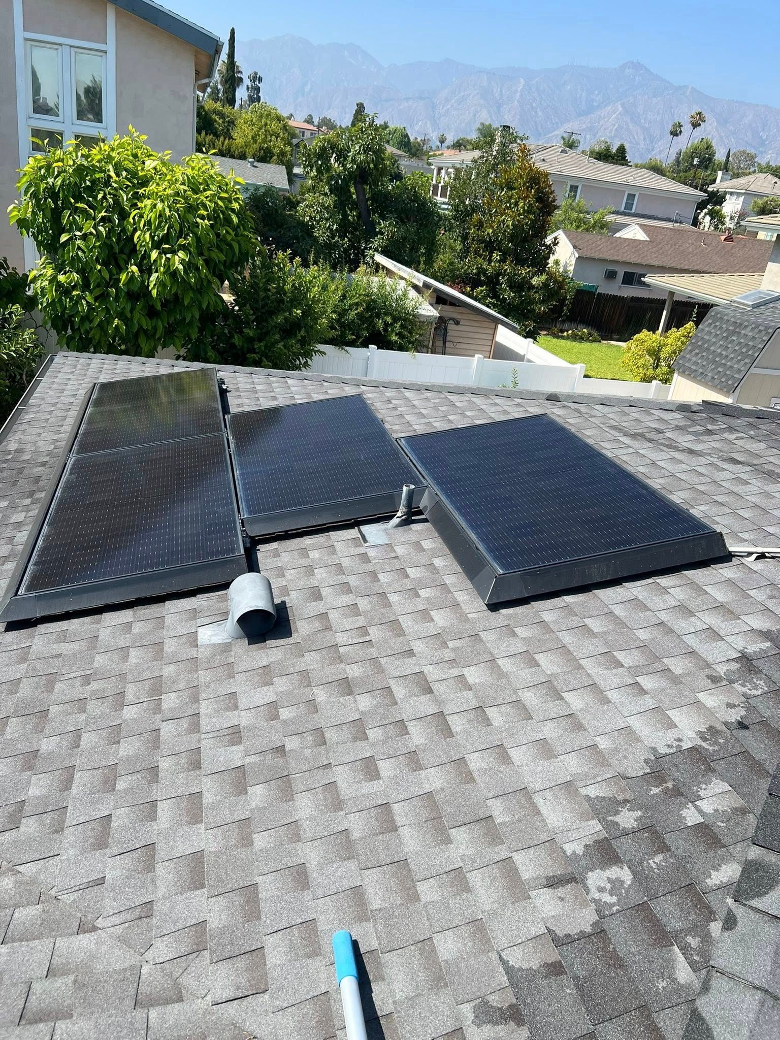 Three black solar panels mounted on a shingled residential roof overlooking a neighborhood and distant mountains.