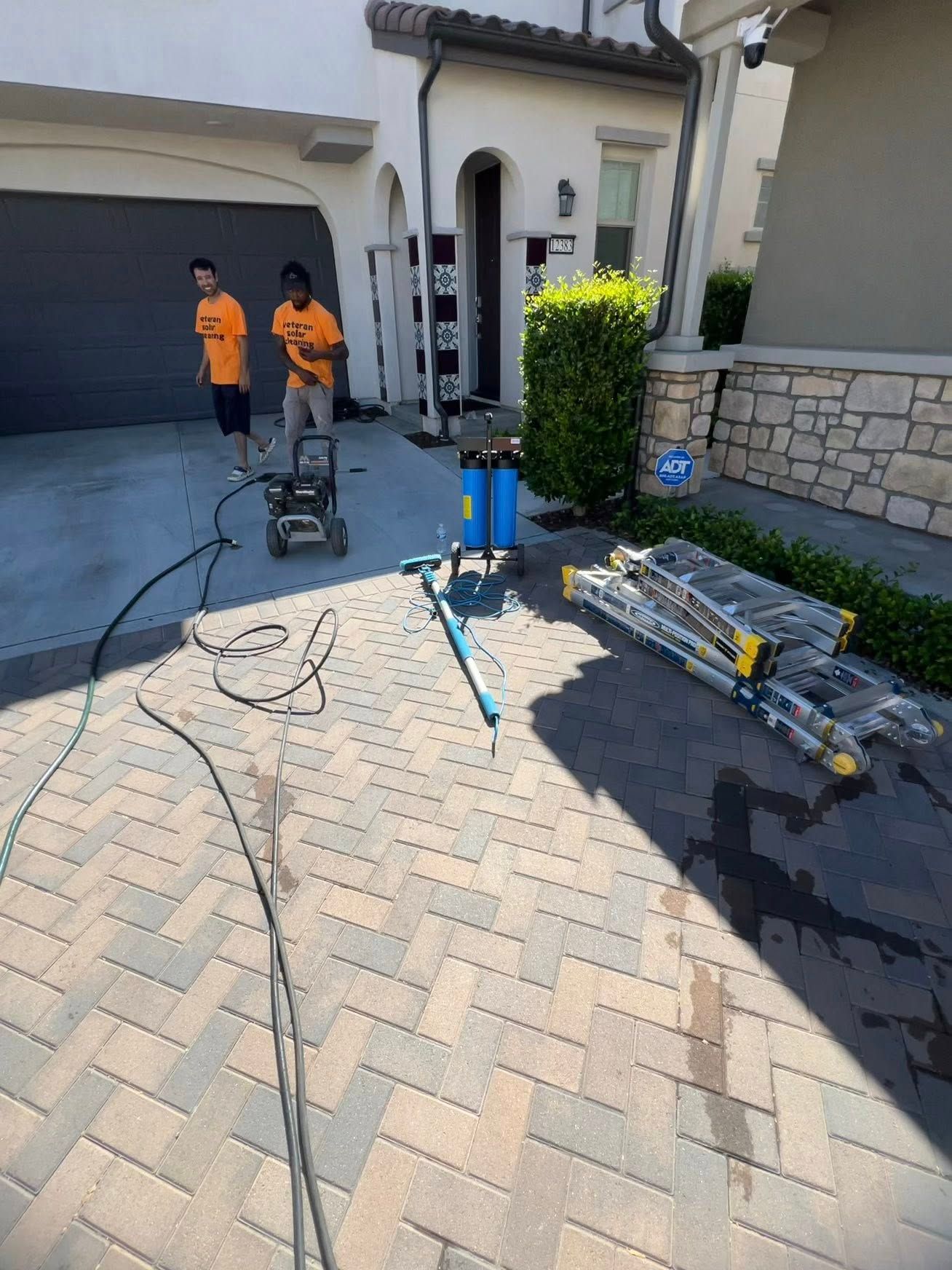 Two workers in orange shirts operate pressure-washing equipment on a residential paver driveway.
