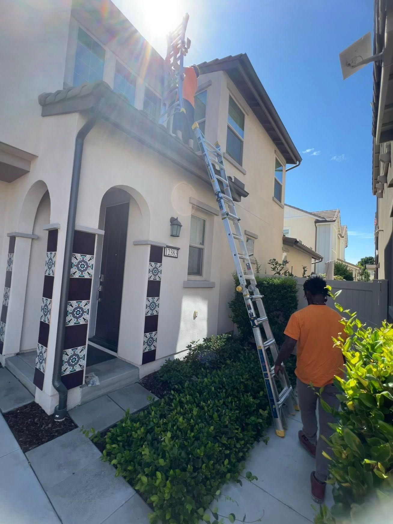 A person in an orange shirt stabilizes a tall ladder leaned against the second story of a tan house.
