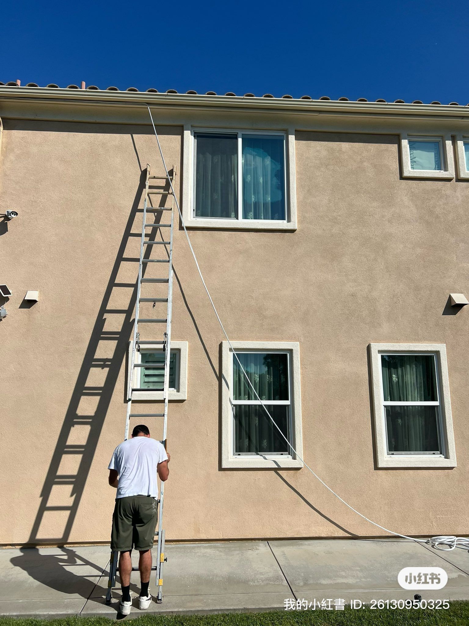 A person in a light shirt and dark shorts stands at the base of an extension ladder leaning against a tan house wall.