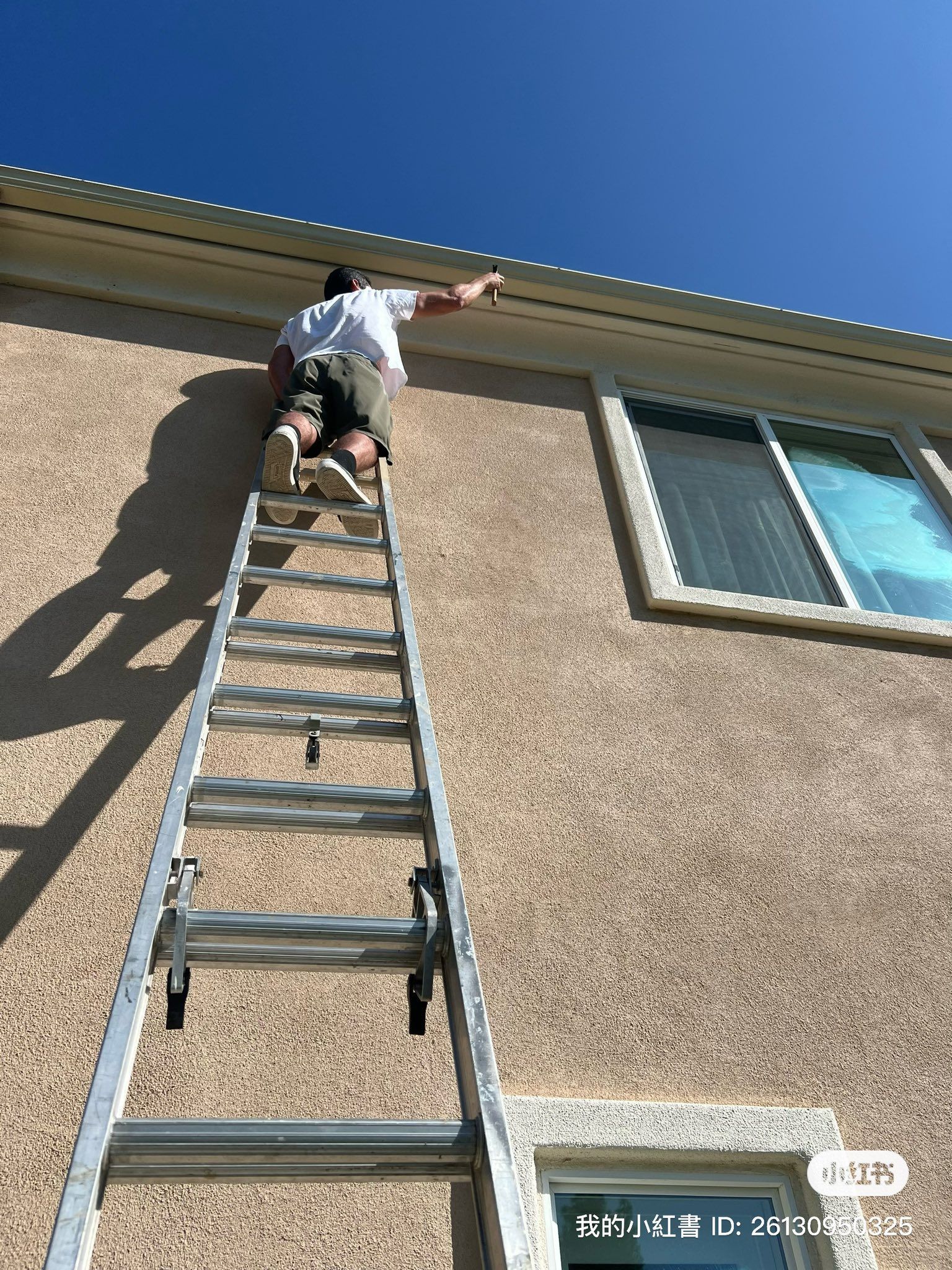 A person standing on an extension ladder leaning against a light-colored stucco house while working on the roofline.