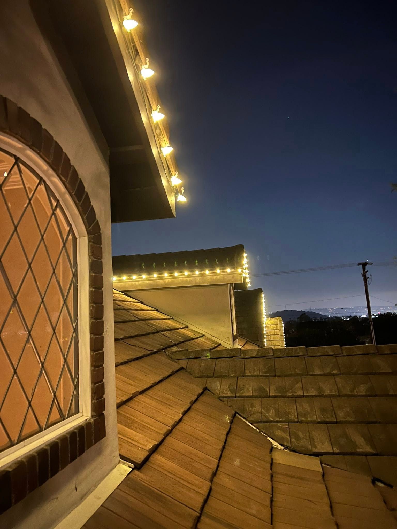 A close-up view of a house roof with warm decorative string lights along the edge against a twilight sky.