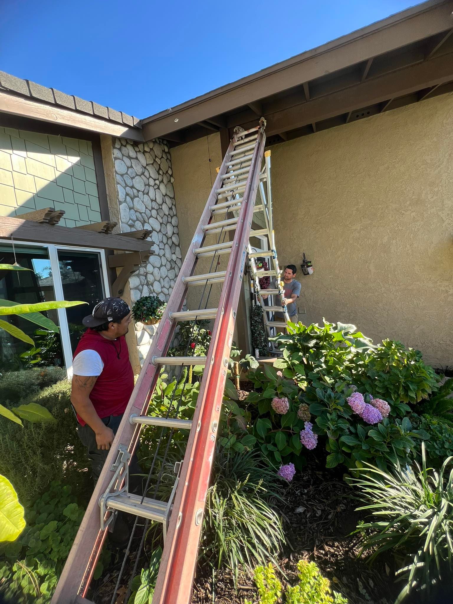 Two people work outdoors at a home, one stands on a tall ladder reaching toward the roof while the other watches below.
