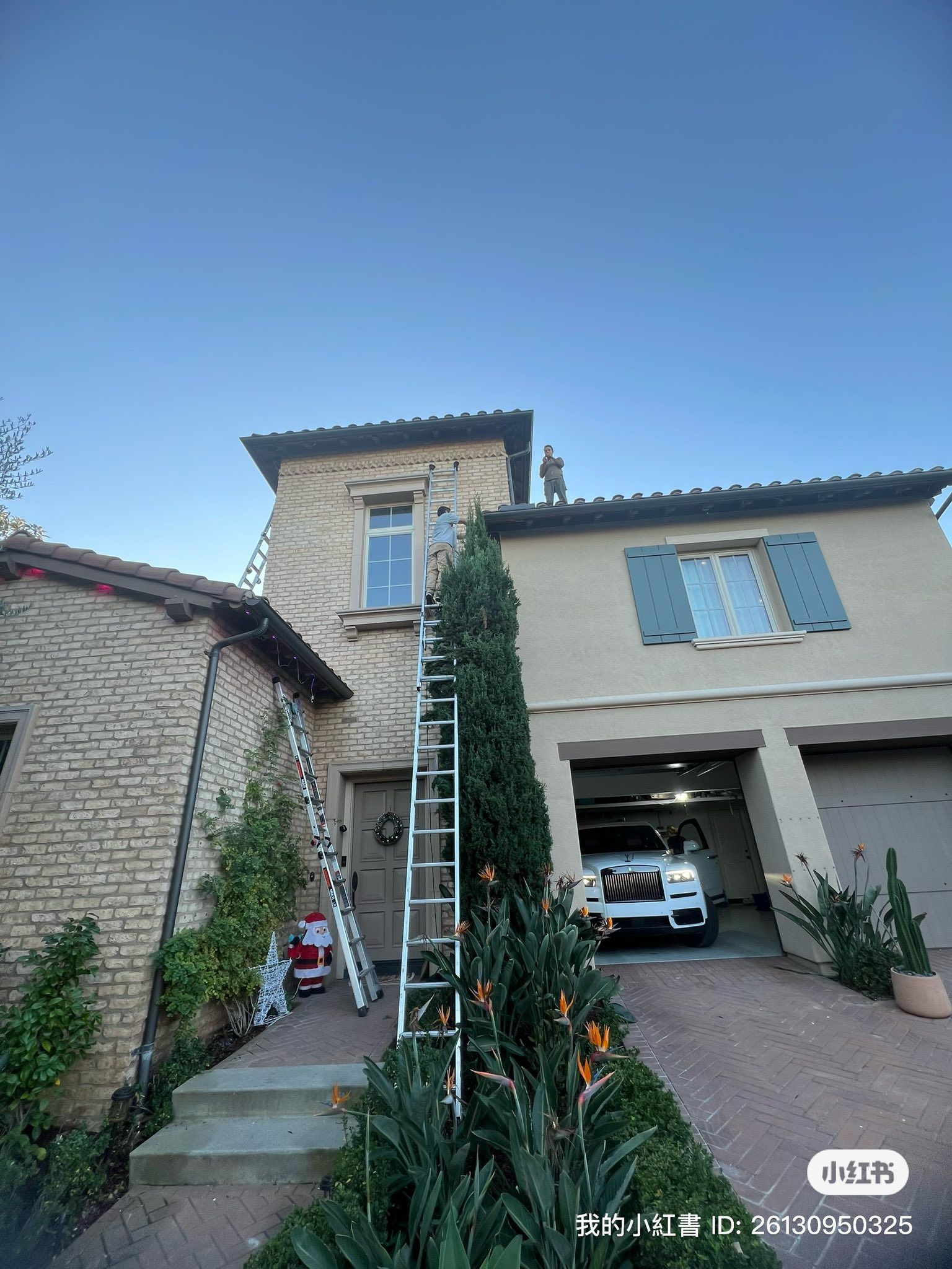 A person on a tall ladder leans against the exterior of a two-story home to hang holiday decorations.