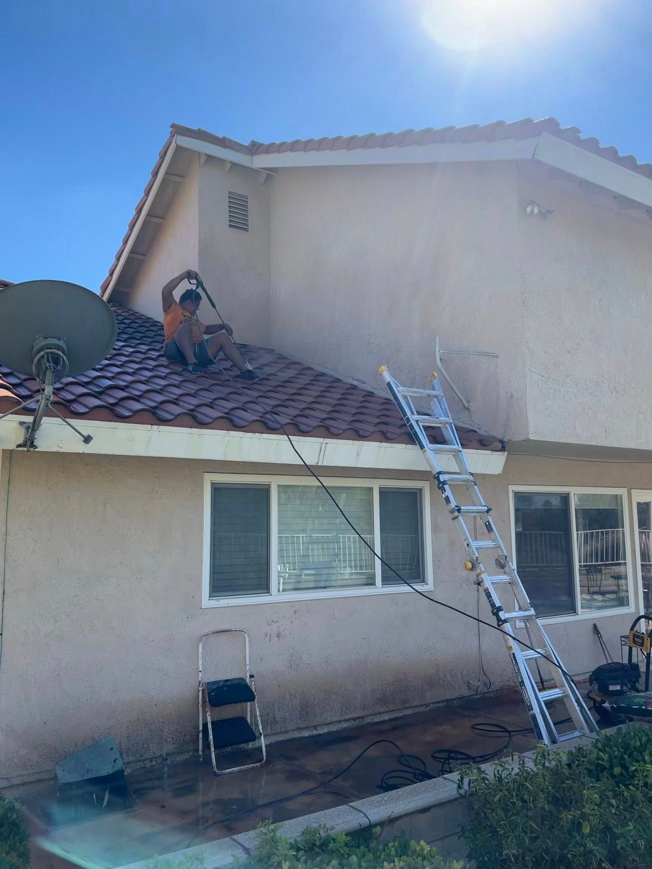A person sits on a tiled roof, using a long-handled tool to wash the side of a tan stucco house next to a tall ladder.