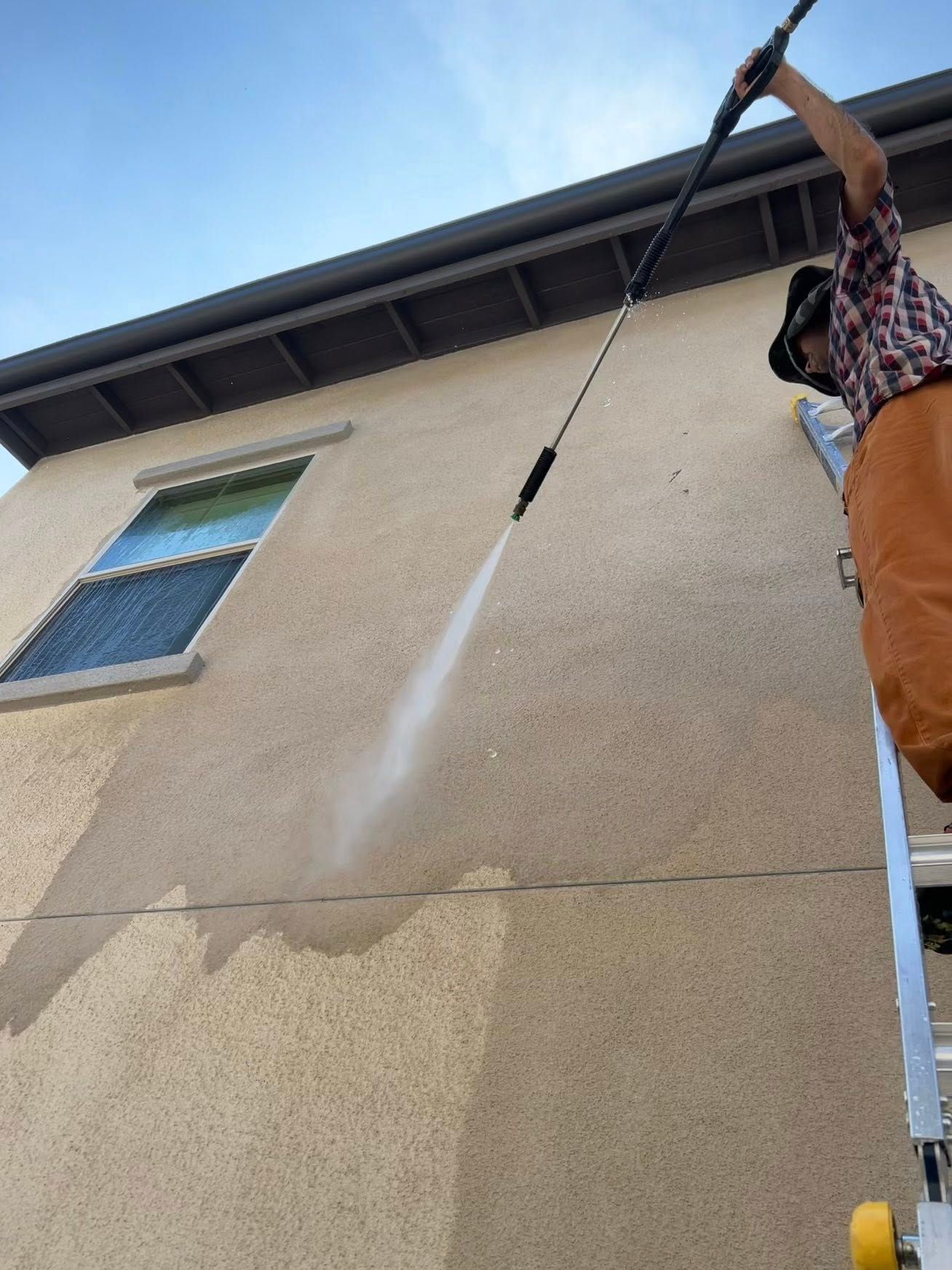 A person stands on a tall ladder, using a pressure washer to clean the light-colored stucco exterior of a building.