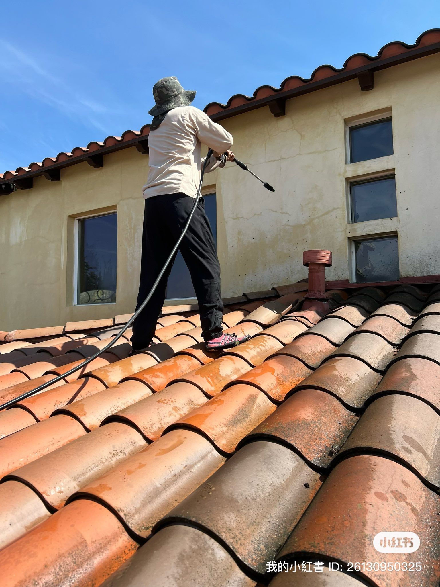A person on a tiled roof uses a pressure washer to clean the exterior wall of a building under a clear blue sky.