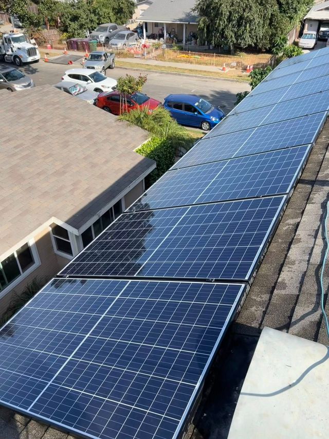 A row of dark solar panels mounted on a slanted residential rooftop, overlooking a neighborhood street with parked cars.