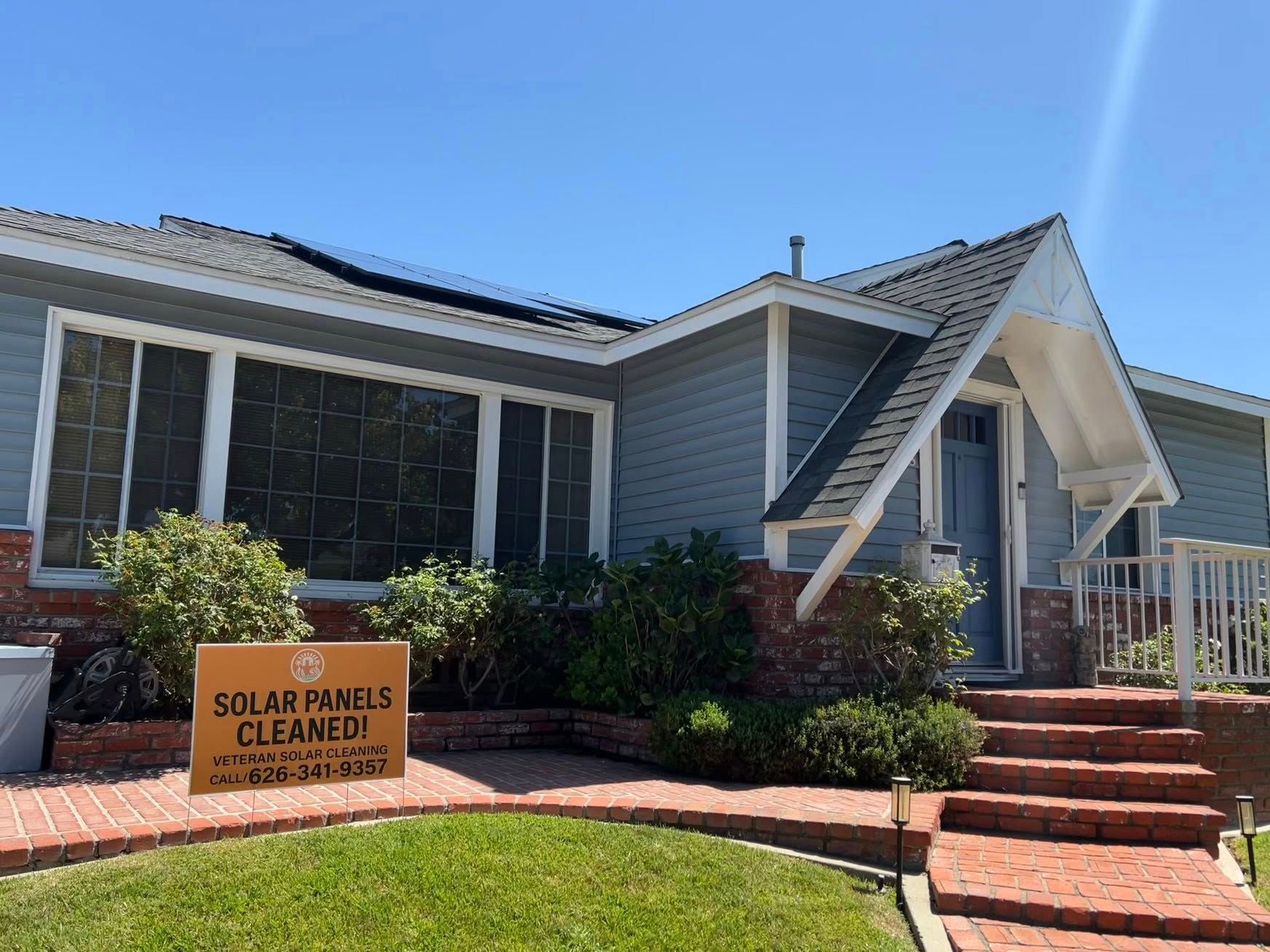 A one-story gray house with a brick entryway and a sign on the lawn that reads 