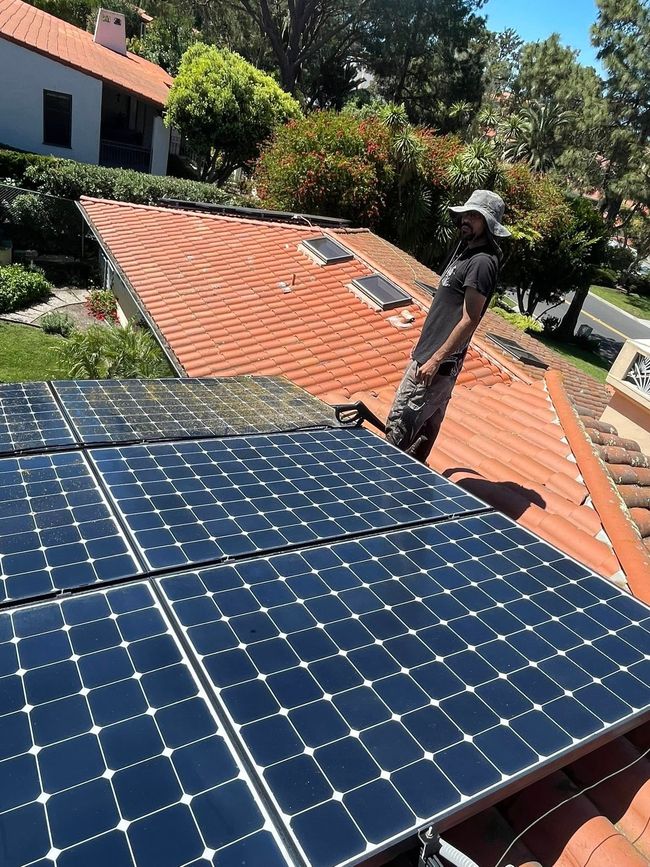 A person in a sun hat stands on a sloped terracotta tile roof next to a newly installed rectangular solar panel array.