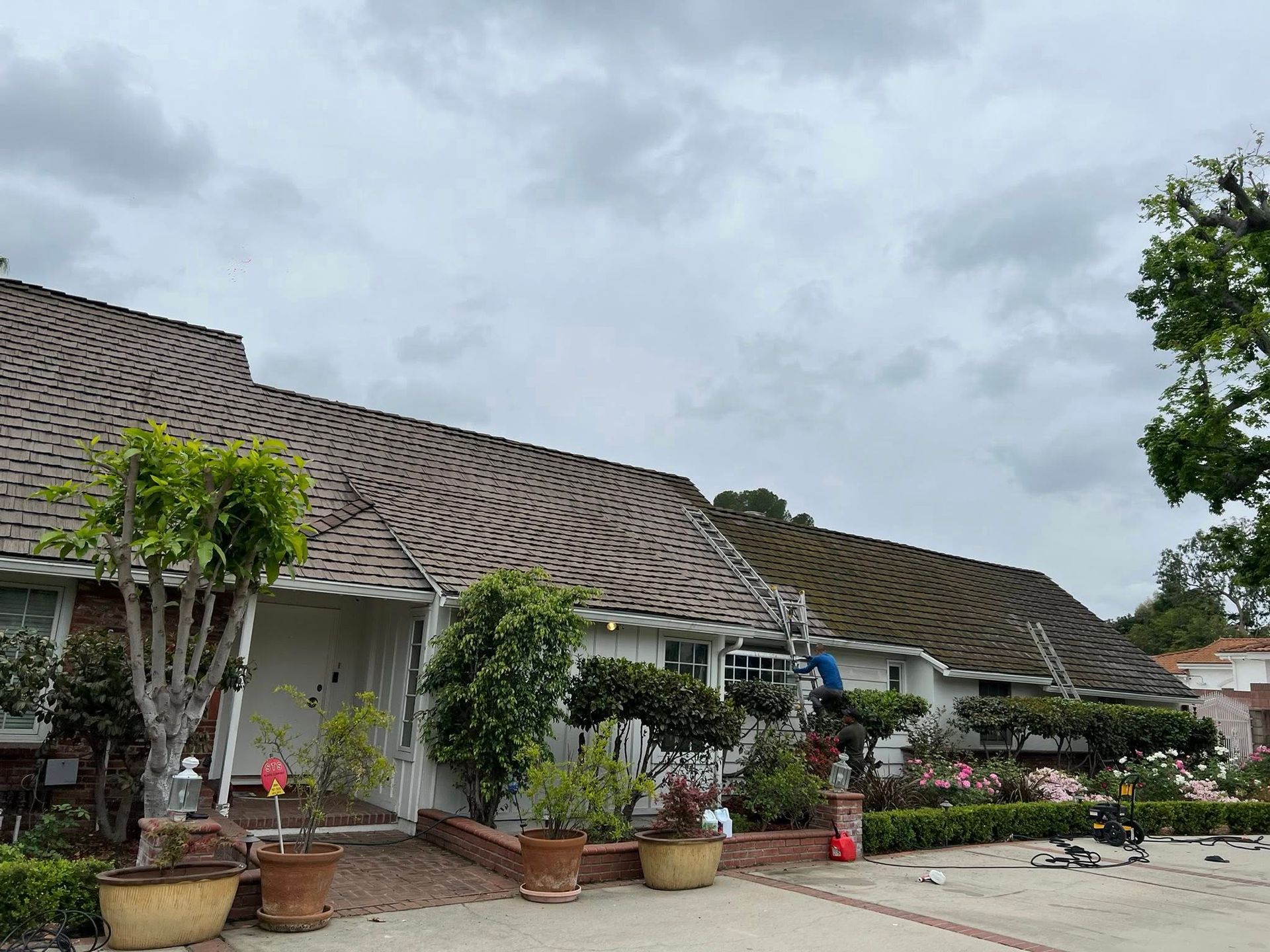 A person on a ladder works on the roof of a one-story, light-colored house with a textured gray roof under a cloudy sky.