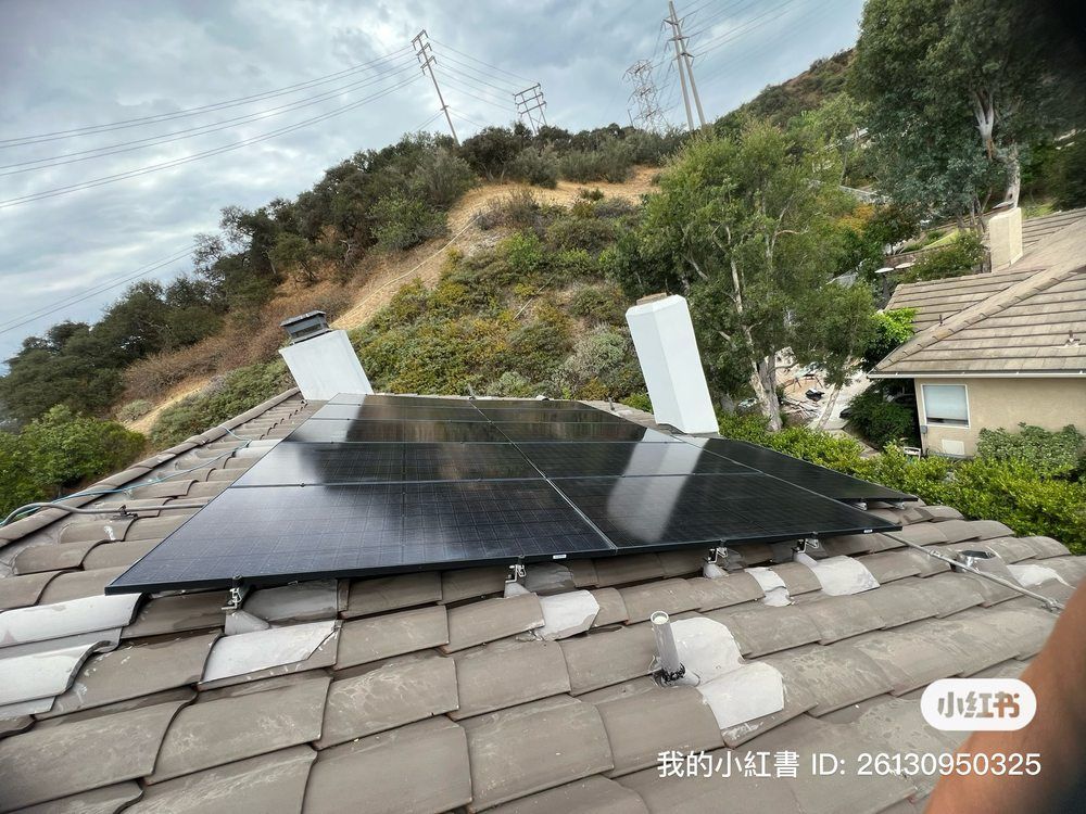 A solar panel array installed on a sloped, gray tile roof, positioned between two white chimneys against a hillside backdrop.