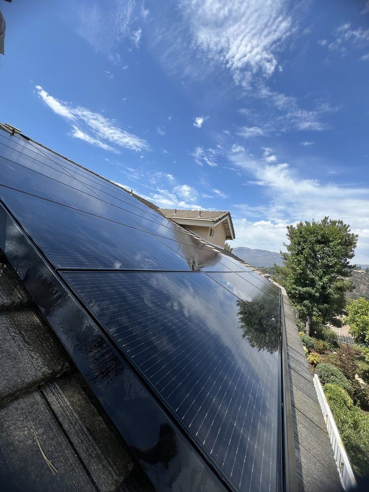 Solar panels on a shingled roof under a bright blue sky with wispy clouds, overlooking trees and distant hills.