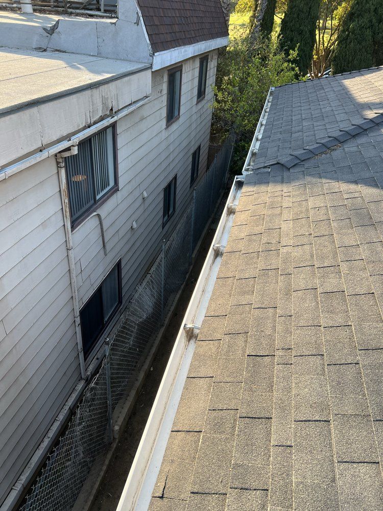 A view looking down into the narrow, fenced gap between the side of a light-colored house and an adjacent shingled roof.