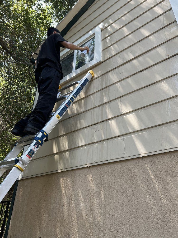 A worker in black clothes stands on a tall ladder to clean a window on the side of a house.