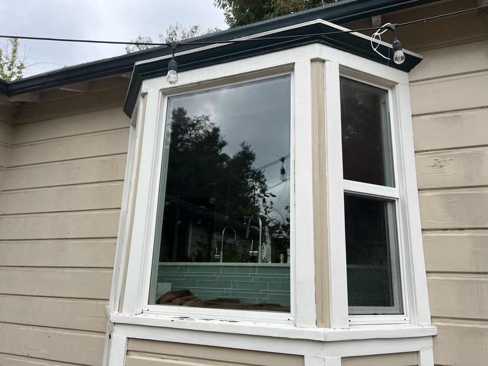 A bay window on a tan, wood-sided house with white trim and hanging patio lights above.