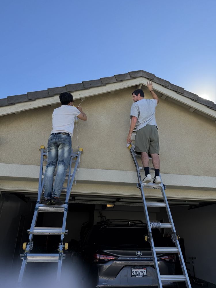 Two people stand on ladders, installing string lights along the edge of a garage roof above a parked car.