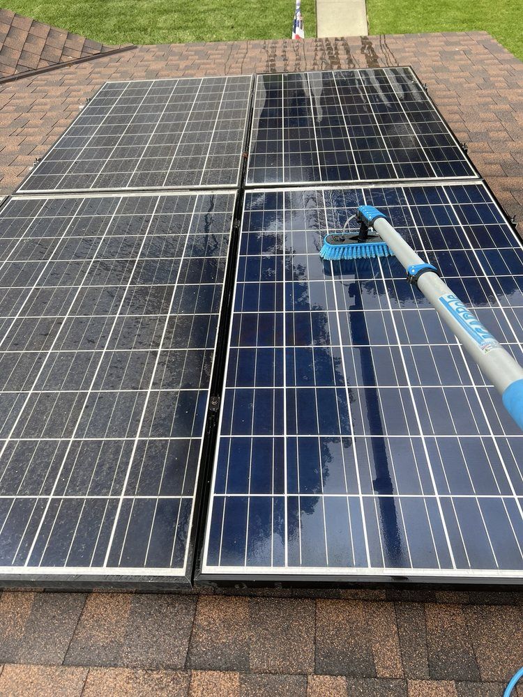 A pole-mounted brush cleans dirt from a section of solar panels mounted on a dark shingled roof.