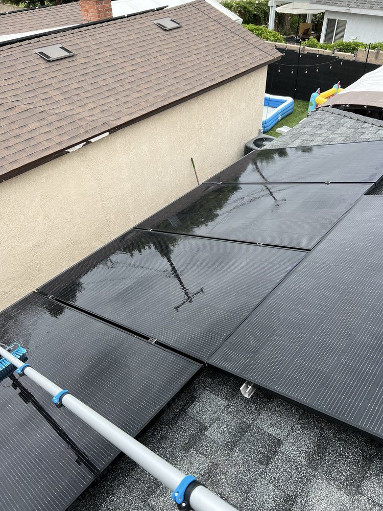 A solar panel array on a shingled roof being cleaned with a long-handled brush tool.