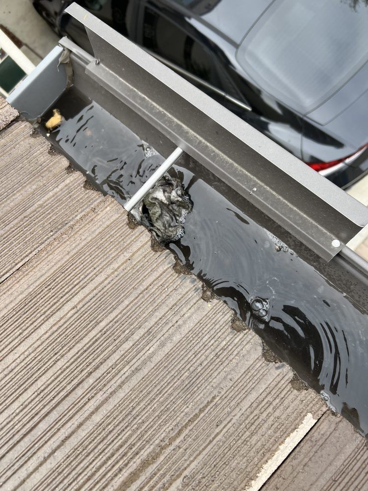 A roof gutter filled with standing water, debris, and sealant patches, seen from a high angle above a parked black car.