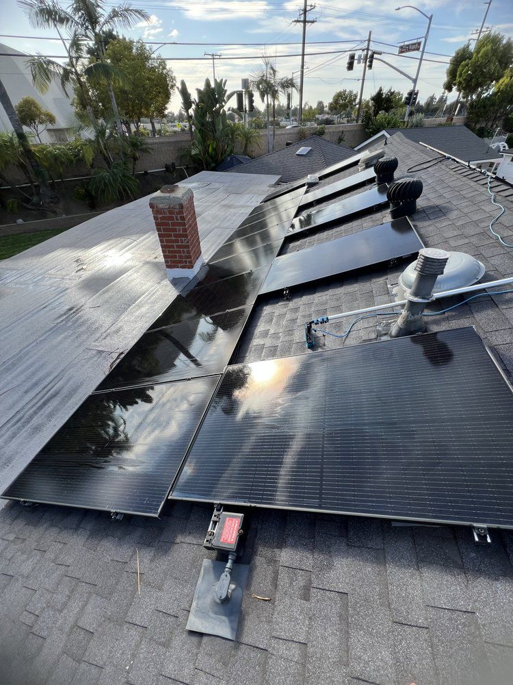 A row of black solar panels mounted on a shingled residential roof next to a brick chimney under a bright sunny sky.
