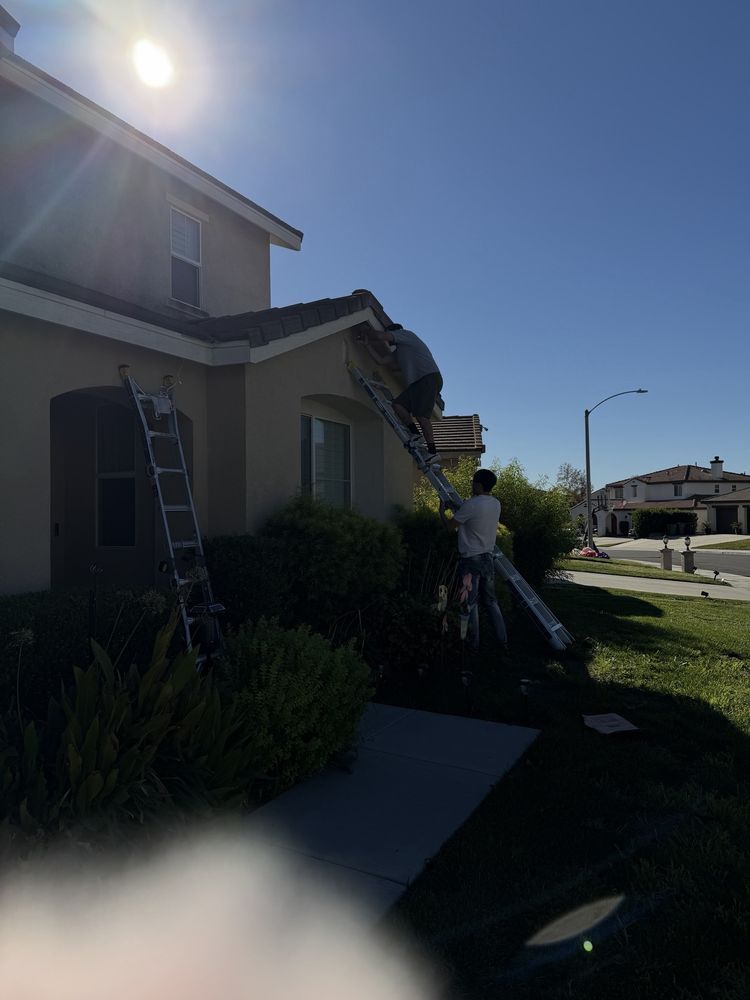 A person on a ladder repairs the roofline of a tan, two-story suburban house on a sunny day.