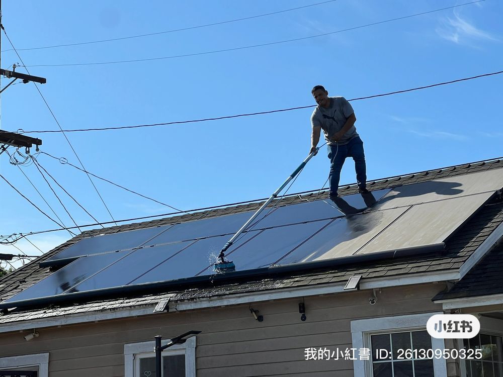 A person stands on a house roof, using a long-handled brush to clean solar panels under a clear blue sky.